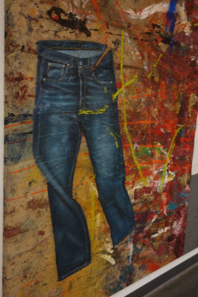 A pair of jeans is painted on a large piece of burlap, with multicolored splatters in the background