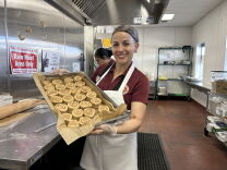 A woman wearing a red polo, apron and hair net holds a baking sheet with freshly rolled cinnamon rolls.
