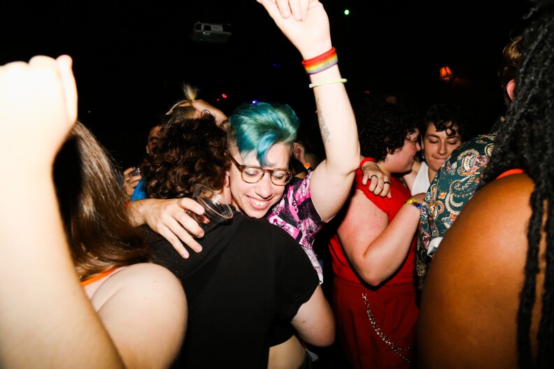 A crowd of people close together in doors. In the center of the photo are two people with light skin tones hugging and smiling. The person facing the camera has blue-green hair, a rainbow wristband and their arm in the air.