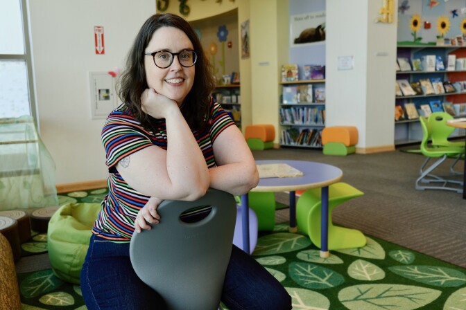 A woman with shoulder-length brown hair wears glasses and a colorful striped short-sleeve shirt. In the background is a green, leaf-patterned rug and bright green chairs. 