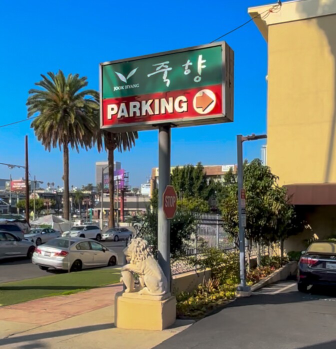 The Jook Hyang parking lot sign with Korean lettering and a red arrow sits under a clear blue Los Angeles sky, flanked by a stone lion statue and surrounded by K-Town’s familiar palm-studded streetscape.