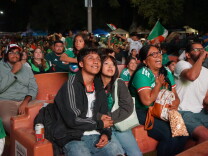 A crowd watches a World Cup match on a large outdoor screen. Many of them wear green jerseys. A young man and woman hold hands, sitting on a bench.