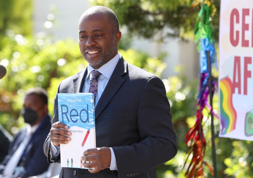 Tony Thurmond wears a suit and smiles as he poses holding up a book titled "Red."