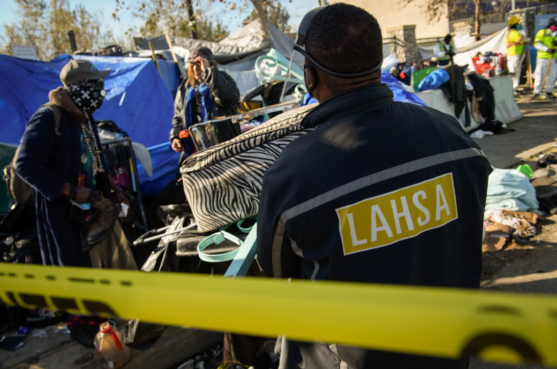 Three people in black jackets stand outside near yellow police tape and a tent being used as shelter. One person wears a jacket with the letters "LAHSA."