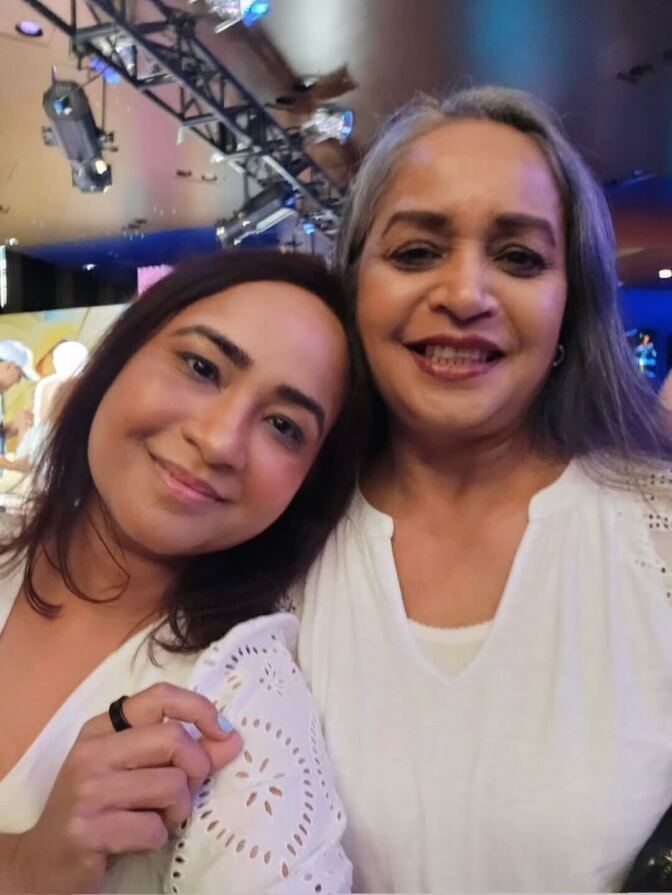 Two South Asian women wearing white lean on each other smiling. 