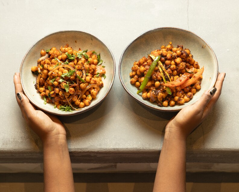 Two bowls of seasoned, cooked chickpeas and vegetables being by a pair of medium-toned hands.
