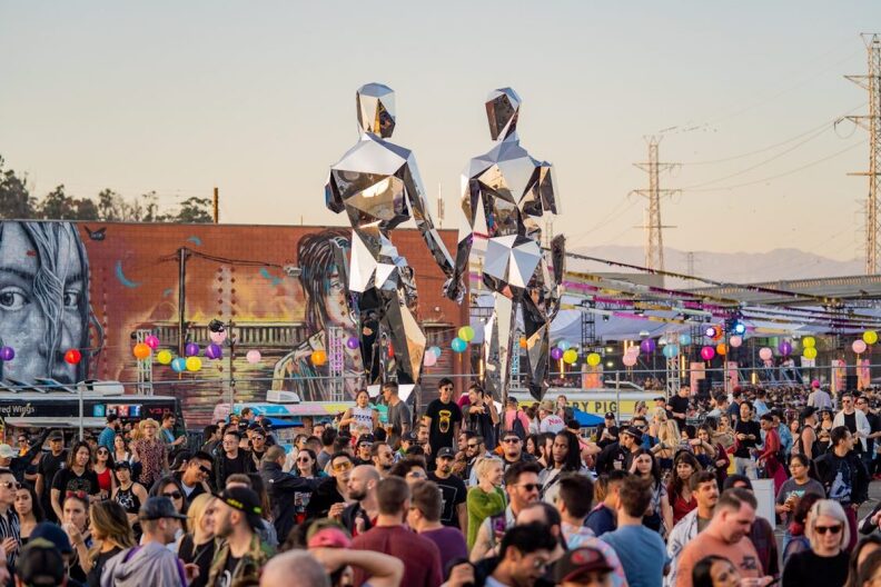 two giant, angular, silver metal statues shaped like humans rise above a crowd of people milling about