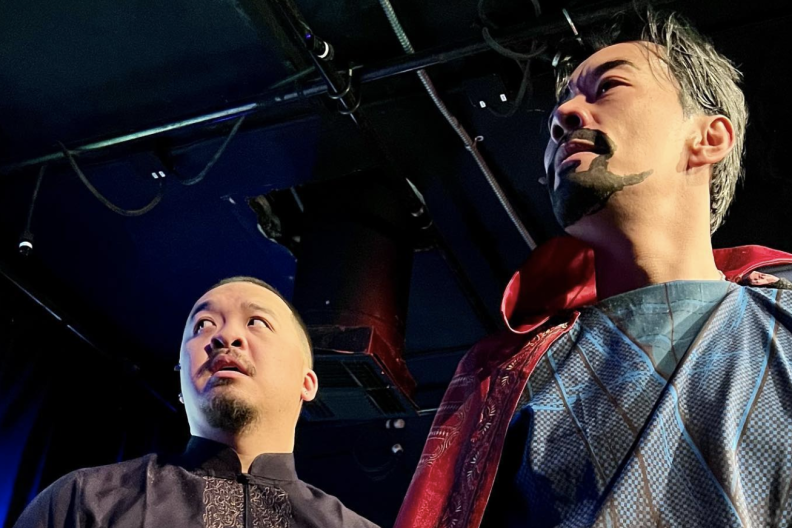 An actor dressed in an Asian-styled to at the left with a surprised look on his face, while the actor on the right is dressed like Marvel superhero Doctor Strange with a blue textured shirt and red cape, with a drawn-on goatee on his face, and a determined look. Stage scaffolding is above them.