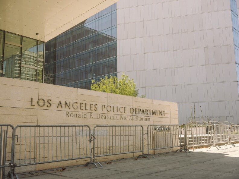 A tan brick wall with letters on it for the Los Angeles police department at a building. There is a silver fence in front.