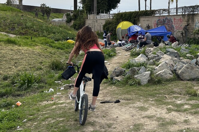  In the foreground, a woman is riding a bicycle away from the camera. In the background, a group of people sit around a tent and some blue tarps between a graffitied and razor wire-topped brick wall, on the right, and, on the left, a drainage ditch. 