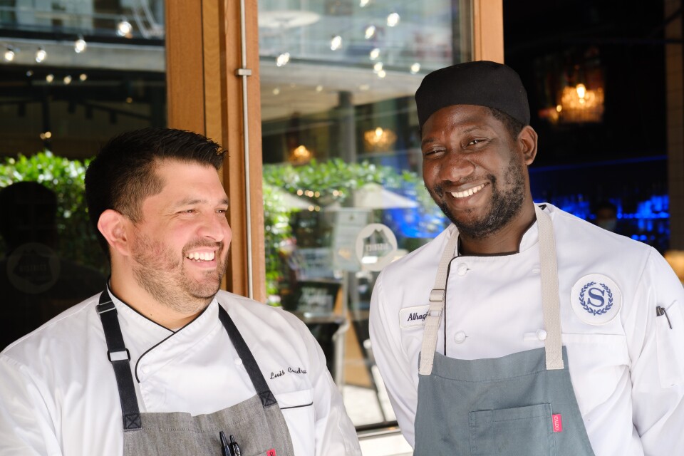 Two men wearing aprons stand next to each other. Both men are smiling and the one on the right looks at the camera. We see a logo with the letter S on the shoulder of the man on the right.
