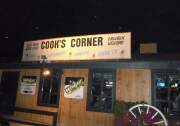 A view of Cook's Corner from the outside. It's one-story building with walls made of vertial brown wooden panels. There is a wood wheel outside by a bush and a big black and and white sign on the top that says, cold beer good food children welcome. Cook's corner.