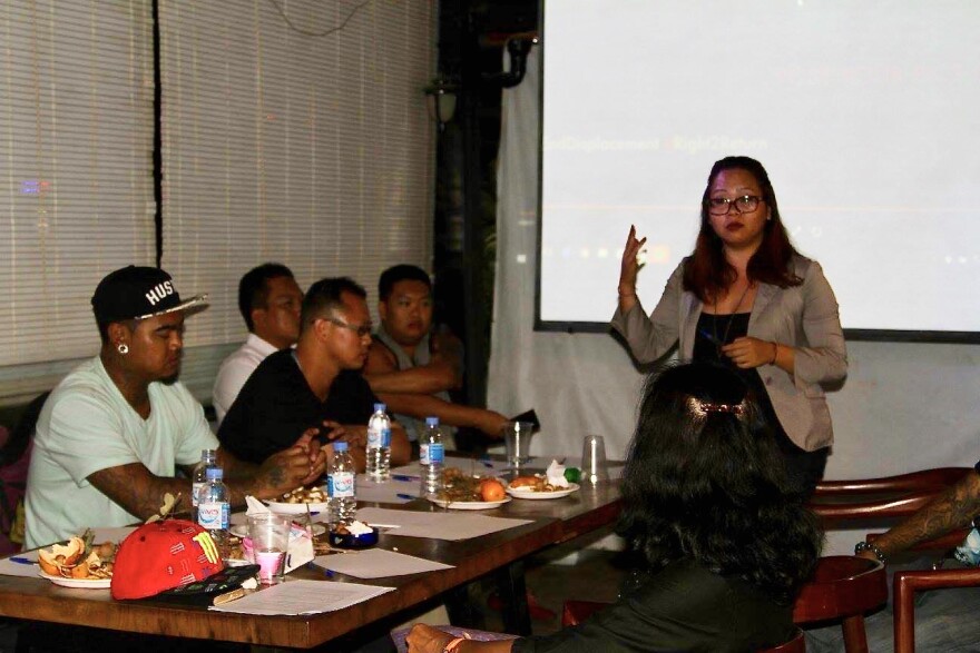 A woman of Asian descent wearing glasses leads a meeting. She stands in front of a projector screen and next to a table where four men are seated.