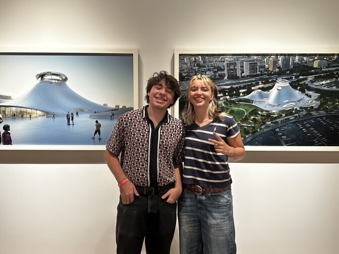 Two people smile together in a gallery, standing before large photos of a saucer-shaped museum building.