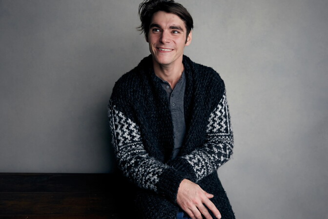 RJ Mitte, a smiling person with dark, tousled hair, sits against a gray studio wall, looking off to the side. They wear a black-and-white patterned knit cardigan over a gray henley.