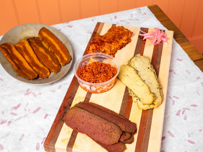 On a striped cutting board an array of offerings, which bacon-looking strips on a nearby plate.