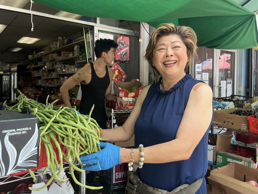 A smiling Asian woman stands at the entrance of a small market under a green tarp,  holding a bunch of long green beans. Behind her, a younger Asian man stands amid stacked produce boxes.