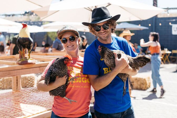 A woman with light skin tone in a red shirt and sunglasses and a man with light skin tone in a blue shirt and sunglasses each hold a chicken.