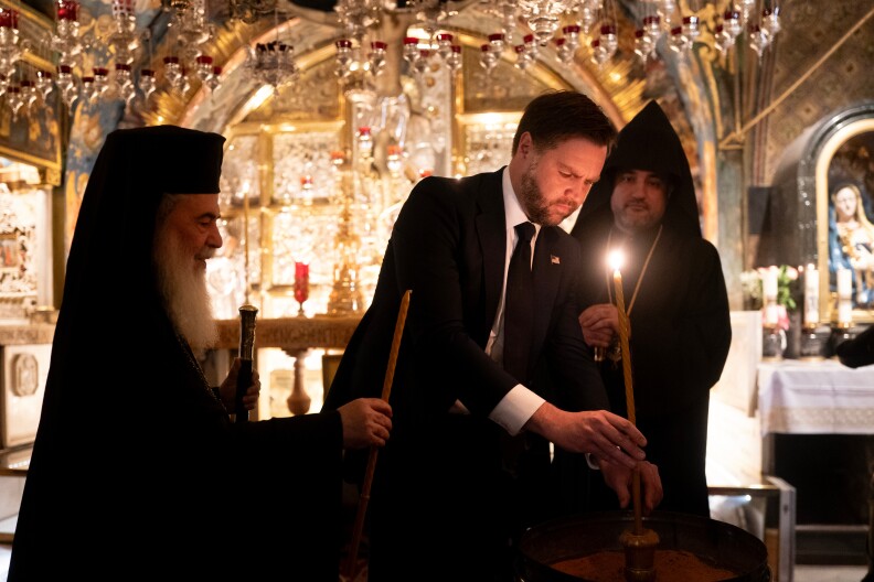 A man in a suit lights a candle in a church accompanied by men in robes.