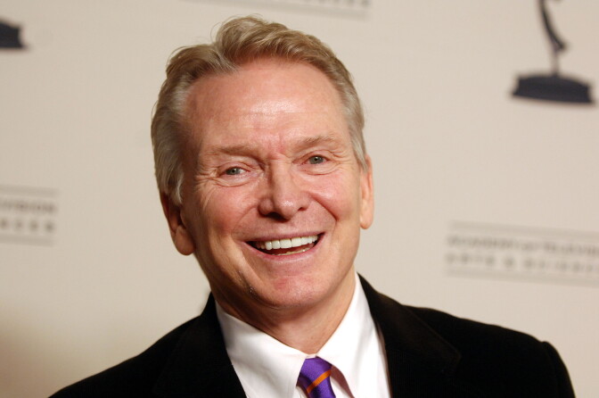 Close up of a smiling man wearing a black suit and purple tie. He is standing in front of a white wall with blurred letters.