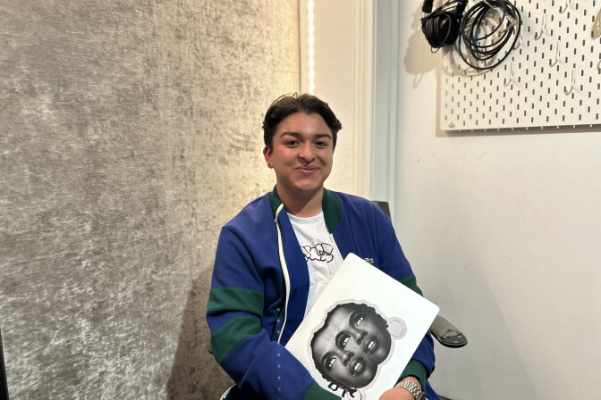 A young person with light brown skin, short dark hair smiles for the camera. They wear a blue sweater and hold a computer with a large sticker of a woman's face in black and white. 