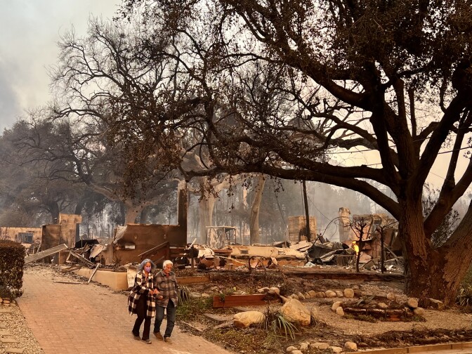 A woman leads a man down a driveway. Behind them is the burned rubble of a home that burned in Altadena due to the Eaton Fire. 