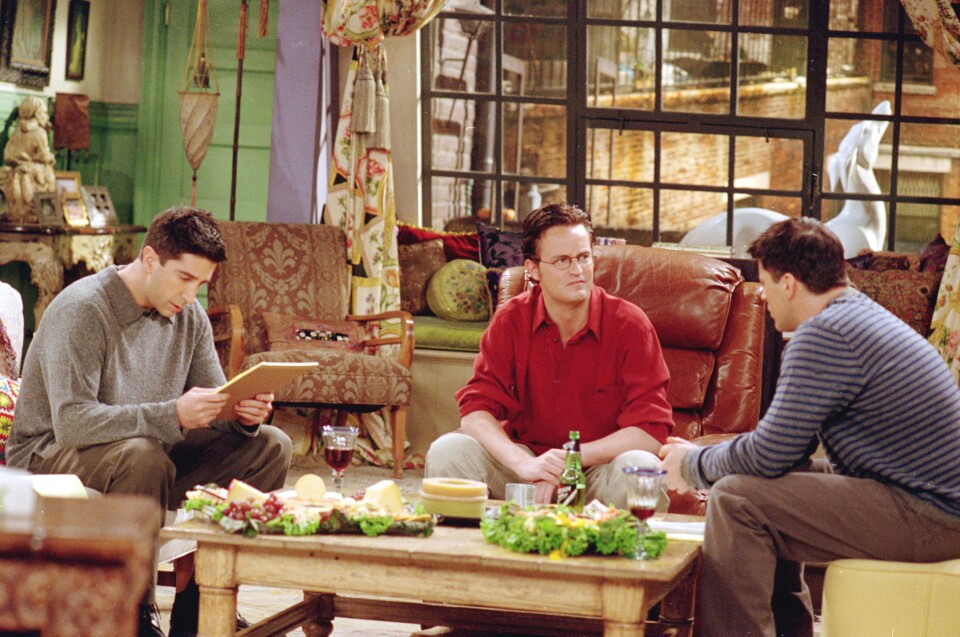 Three men sit around a wooden coffee table with various small bite appetizers on top of it. 