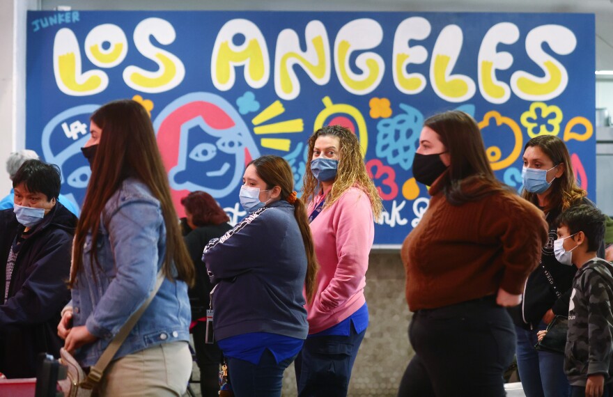 A group of people wearing medical face masks stand in a line. A woman in the center looks at the camera. There's a colorful art piece in the background that says "Los Angeles."