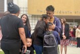 An adult with medium skin tone embraces two children outside a school. Adults and children mix nearby and in the background.