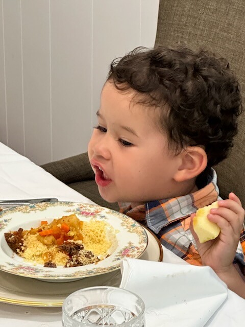 A boy with light brown skin and brown hair sits at a table with a plate of yellow couscous with vegetables on top. In one hand he's holding a cut piece of an apple with a bitemark. 