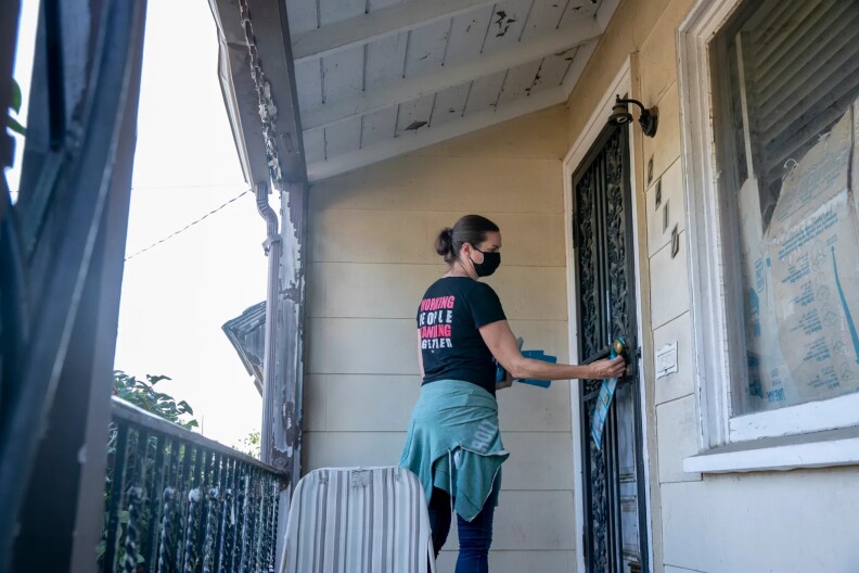 A young woman with light skin and wearing a mask leaves an election handout on a doorknob.