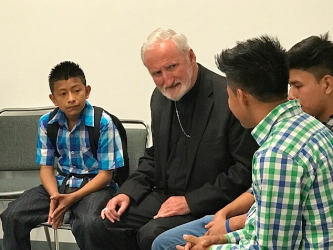 A man in all black with gray hair and a beard sits next to children with medium-tone skin in plaid shirts. One wears a backpack.