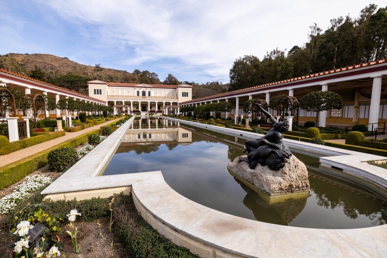 A scenic building and courtyard on a slightly cloudy day. The water in a pool along the center of the space is dark and murky.
