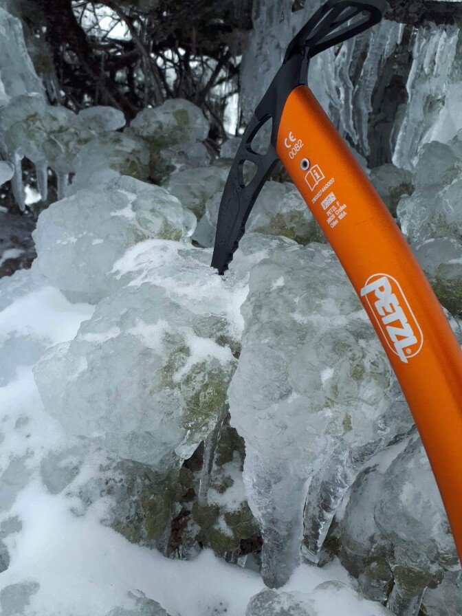 A closeup of an ice ax with an orange handle sticking into the ice covering a tree branch.