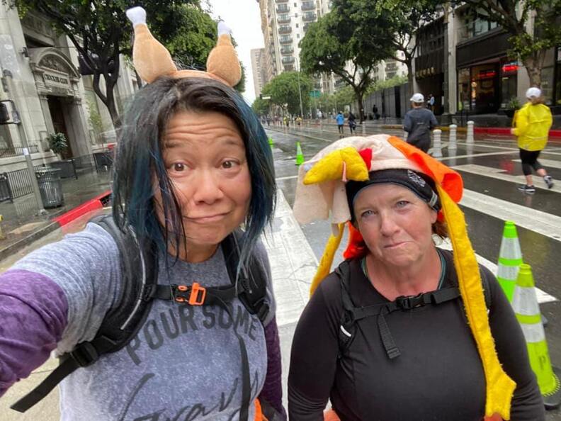 Two women stand with mock sad faces in a rainy Downtown LA street. One is wearing fake turkey drumsticks on her head; the other is wearing a turkey hat with long yellow legs hanging down by her face.