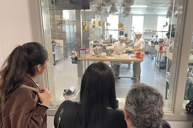 Three family members watch the student chefs work through a large window, where a cooking lab can be seen.