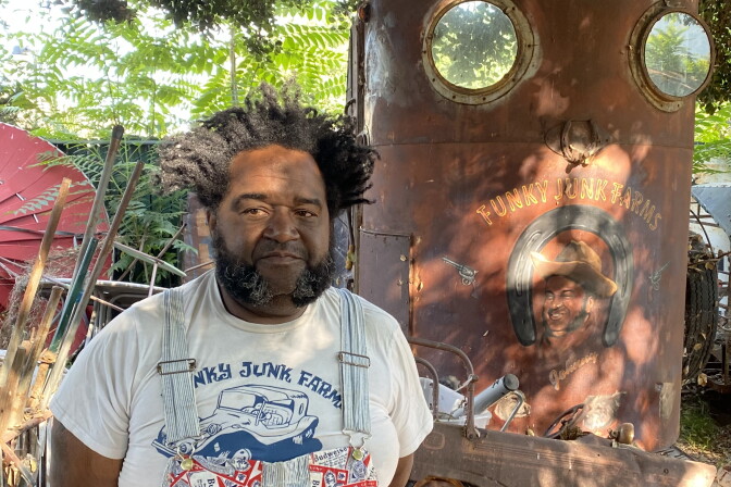 Johnny Agnew of Funky Junk Farms wears overalls and stands in front of a vintage trailer 