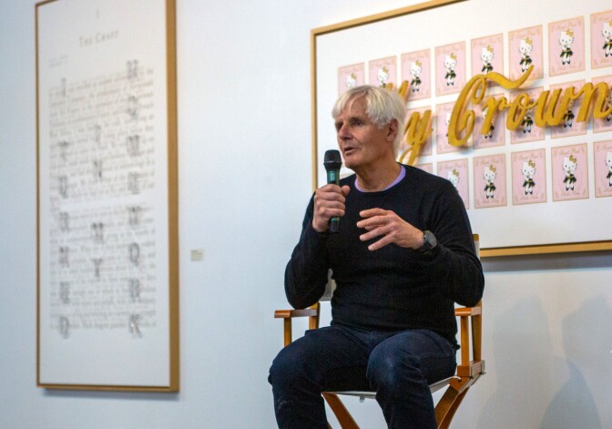 A man with grey hair sits in a chair and speaks while holding a microphone in his right hand. He sits in front of a white wall featuring framed art pieces.