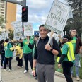 A man with medium skin tone and short dark hair smiles holds up a picket sign. He is clad in gray slacks and a black shirt. Behind him, his co-workers also hold up signs.