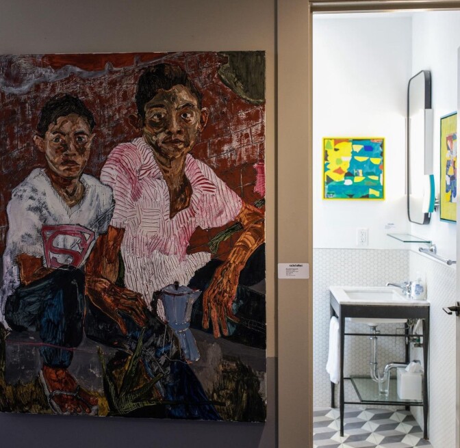 A painting of two children, one wearing a pink shirt and the other a superman shirt, rests on a wall outside a restroom featuring more art.