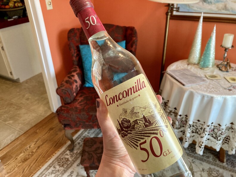 A hand holds a glass bottle with a pale yellow label with red lettering with a detail drawing of field. The cap of the bottle is also red