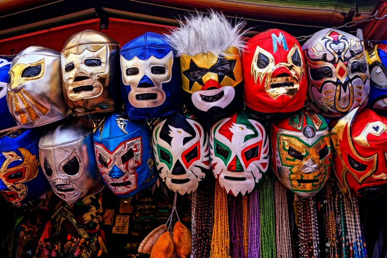 The image is 2 rows of different color/variations of Lucha Libre masks hanging on a rod outside a street vendor.