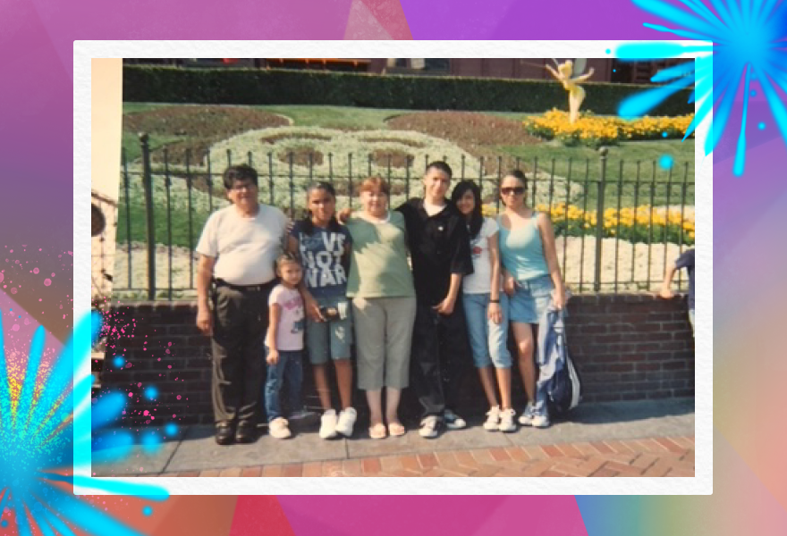 The family of author Jorge Cruz, third from right in a black shirt, at Disneyland circa 2005: His grandfather, grandmother, aunt, young sister and two cousins pose against a landscaped image of Mickey Mouse in greenery and yellow flowers.