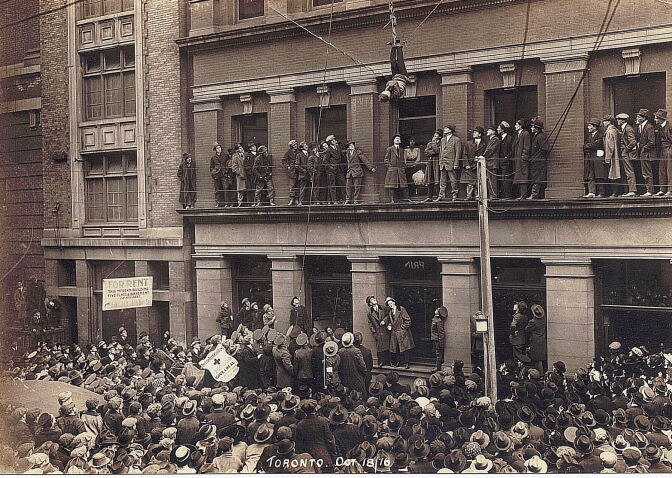 A black and white image shows a crowd in front of a building. A man hangs from his feet two stories up, his arms bound to his body.