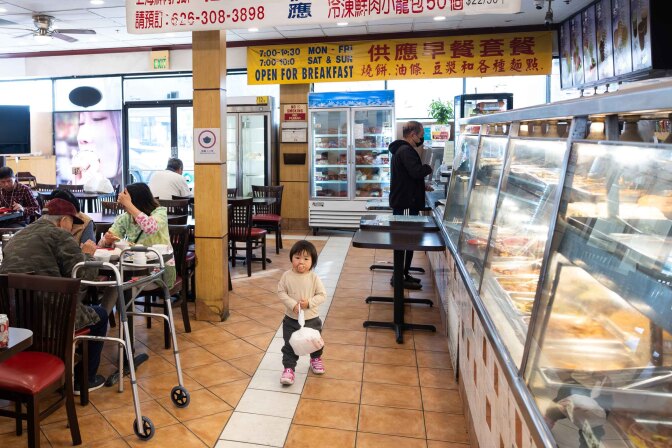 a wide shot of a food court with people sitting at tables on the left, a cute young child standing in the middle with a pacifier in their mouth staring at the camera, and a covered glass display showing many food dishes in stainless steel pans on the right