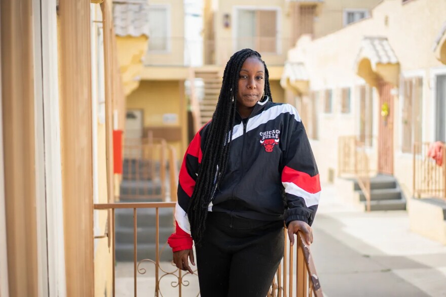 A Black person wearing a red, black and white Chicago Bulls jacket leans against of railing outside and looks to the camera for a portrait. They have long dark hair in braids.