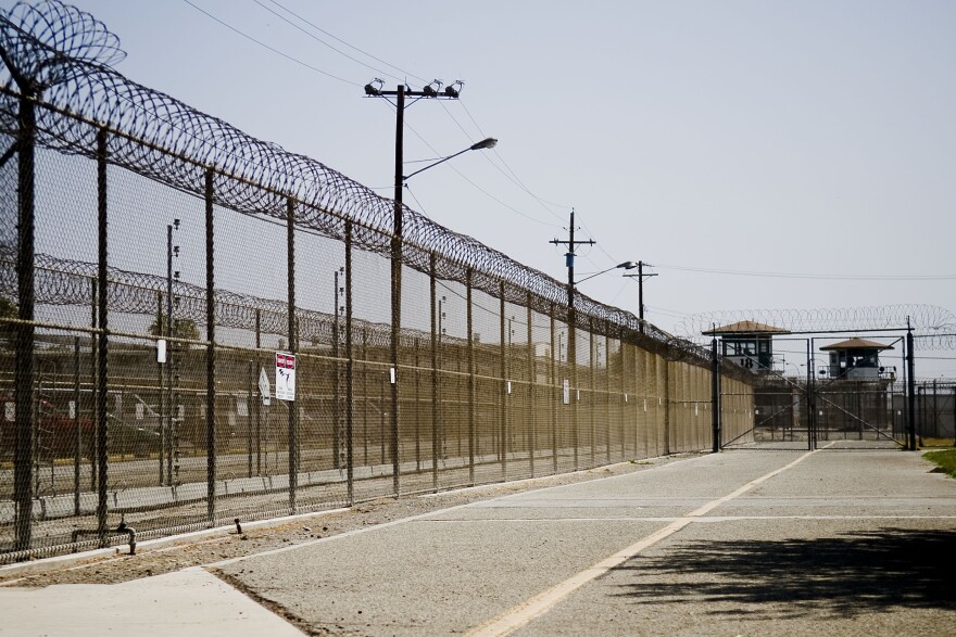 Two prison watch towers stand in the distance behind a long fence topped with barbed wire. Along one stretch of fence is a small black and white sign with a red strip across the top and the symbol of a lightning bolt striking a person.