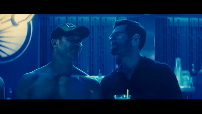 Luke Macfarlane (left) stands next to Billy Eichner (right) in a dimly lit bar that casts a smoky blue hue across the scene. Their shoulders are touching as they look into each other's eyes. Each man has a slight smile, lips closed, as if sharing a private joke.