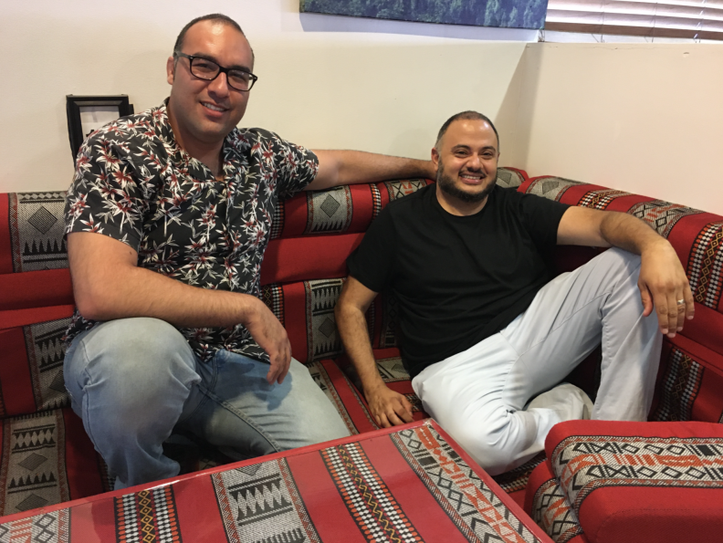 Two men, one in printed shirt, another in a black t-shirt, sit on red cushions against a white wall.