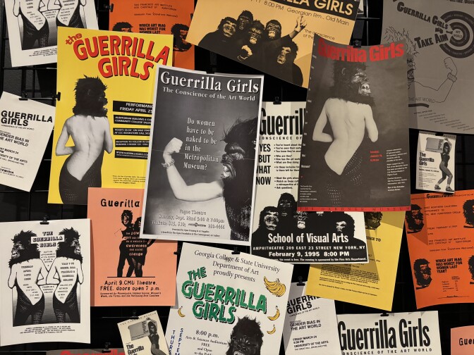 White, orange and yellow flyers pinned to a board with text that includes "Guerrilla Girls" and images of partially nude women wearing gorilla masks.